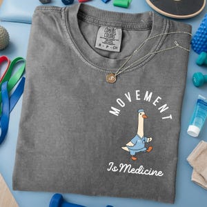 May include: A gray Comfort Colors t-shirt with the words "MOVEMENT Is Medicine" and a cartoon goose wearing a doctor's uniform. A gold necklace with a pendant is on the shirt. Exercise equipment is in the background.