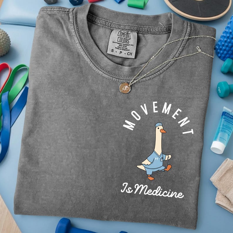 May include: A gray Comfort Colors t-shirt with the words "MOVEMENT Is Medicine" and a cartoon goose wearing a doctor's uniform. A gold necklace with a pendant is on the shirt. Exercise equipment is in the background.