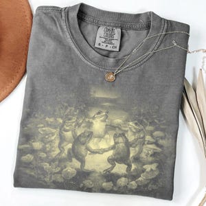 May include: Dark gray Comfort Colors t-shirt featuring a whimsical illustration of dancing frogs in a forest setting. The shirt has a classic crew neck and short sleeves. A silver necklace with a small pendant rests on the shirt. The shirt is folded neatly.