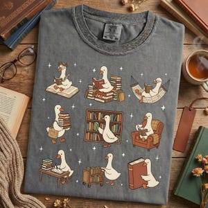 Puede incluir: Camiseta gris con un dise&ntilde;o caprichoso de gansos leyendo libros en varios escenarios. Los gansos est&aacute;n representados con gafas y accesorios, rodeados de libros, estanter&iacute;as y una hamaca. La camiseta tiene cuello redondo cl&aacute;sico.
