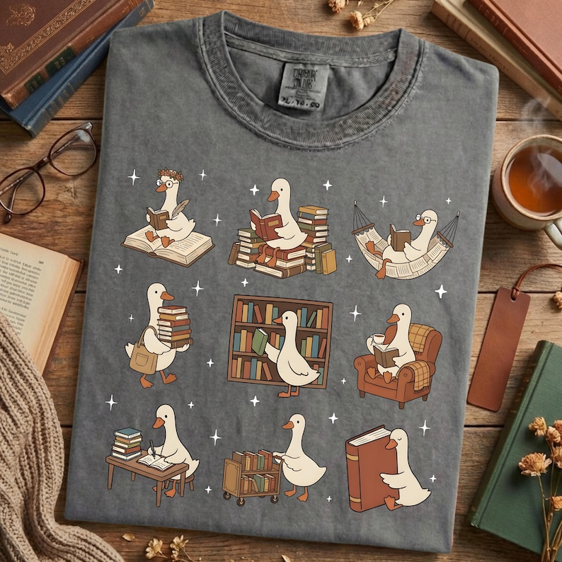 Puede incluir: Camiseta gris con un dise&ntilde;o caprichoso de gansos leyendo libros en varios escenarios. Los gansos est&aacute;n representados con gafas y accesorios, rodeados de libros, estanter&iacute;as y una hamaca. La camiseta tiene cuello redondo cl&aacute;sico.