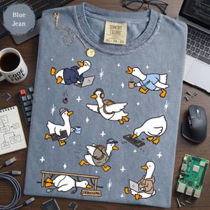 May include: A blue jean colored t-shirt featuring a graphic print of multiple white geese engaged in various computer programming activities, including typing on laptops, wearing headphones, and holding mugs, with small star accents throughout.