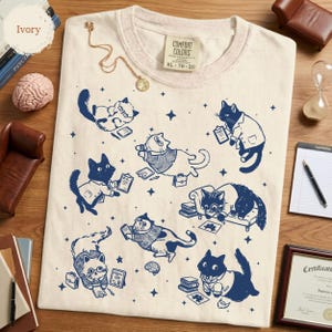 May include: An ivory t-shirt featuring a dark blue graphic print of various cats in professional attire, including lab coats and sweaters, surrounded by small stars, books, clipboards, and briefcases. The shirt has a Comfort Colors brand label.