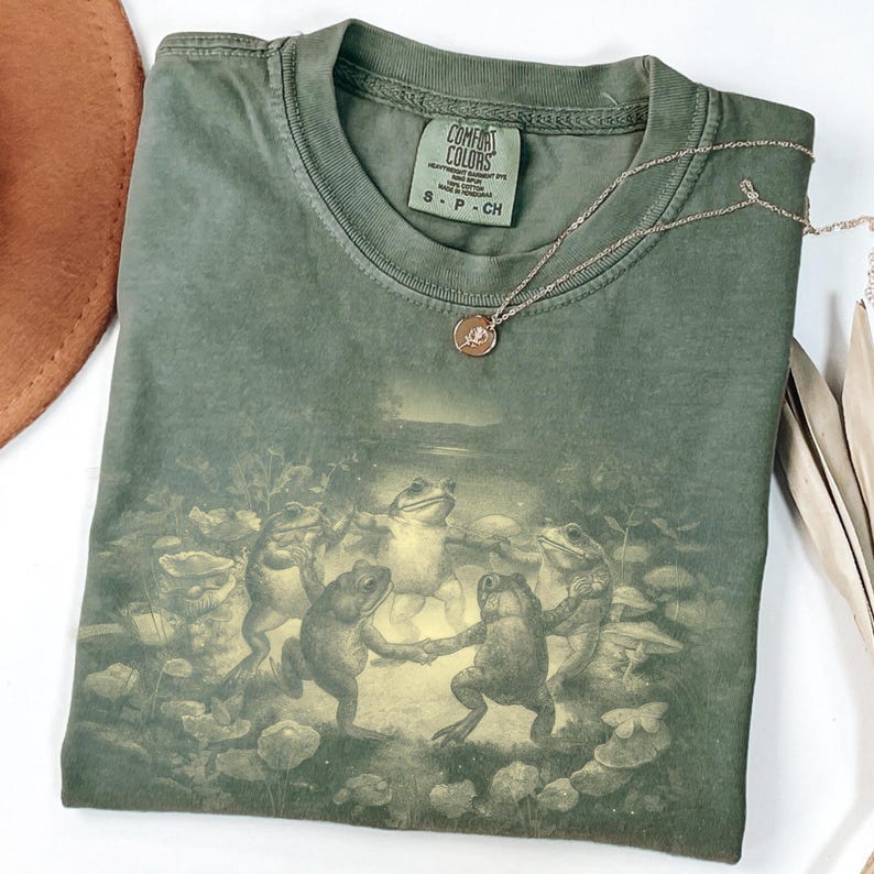 May include: Green Comfort Colors t-shirt featuring a whimsical illustration of frogs dancing in a circle. A delicate silver necklace with a small pendant rests on the shirt. The shirt is folded, with a brown hat visible in the background.