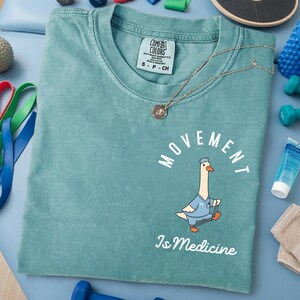 May include: A teal Comfort Colors t-shirt with the words "MOVEMENT Is Medicine" and a cartoon goose in a doctor's uniform. A silver necklace with a small pendant rests on the shirt. The shirt is size small.