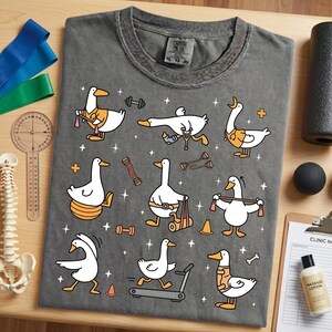 May include: Dark gray t-shirt featuring cartoon ducks engaged in various physical therapy exercises. The ducks are depicted with orange accents and are using weights, treadmills, and resistance bands. The shirt includes the text "Urban Wear" on the tag.