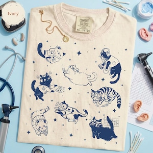 May include: Ivory-colored Comfort Colors t-shirt with a crew neck, featuring navy blue illustrations of cats in various medical and tech-related poses. The shirt has a size tag of XL 78-20. A gold necklace and medical equipment are also visible.