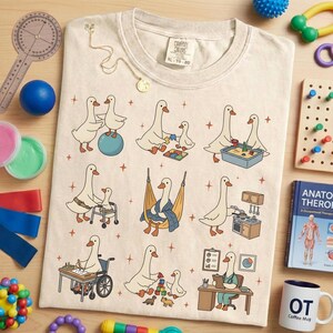 May include: Cream-colored t-shirt with a whimsical design featuring cartoon geese in various occupational therapy scenarios. The shirt includes the text "COMFORT COLORS" and size "XL 70-20". Surrounding the shirt are occupational therapy tools.