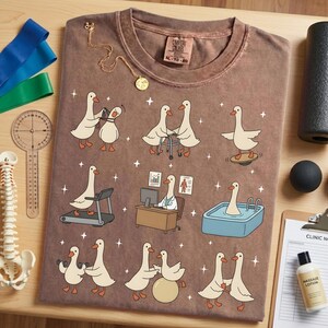Pode incluir: Camiseta marrom com gansos de desenho animado em situa&ccedil;&otilde;es de fisioterapia. A camiseta tem gola redonda e uma etiqueta. Um colar de ouro e equipamentos m&eacute;dicos tamb&eacute;m s&atilde;o vis&iacute;veis. Tamanho XL-TG-2X.