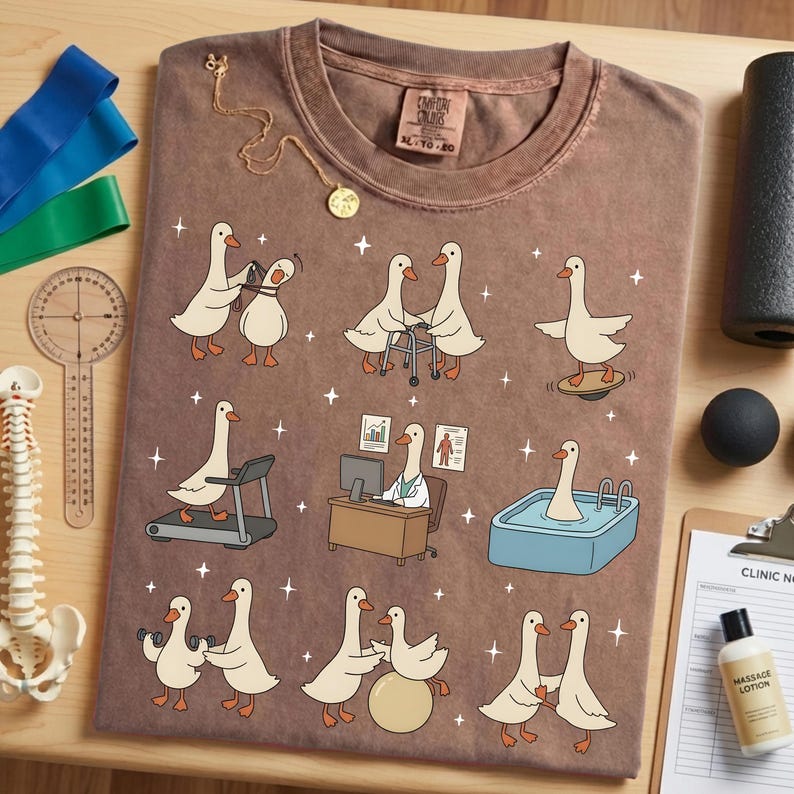 Pode incluir: Camiseta marrom com gansos de desenho animado em situa&ccedil;&otilde;es de fisioterapia. A camiseta tem gola redonda e uma etiqueta. Um colar de ouro e equipamentos m&eacute;dicos tamb&eacute;m s&atilde;o vis&iacute;veis. Tamanho XL-TG-2X.