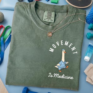 May include: Olive green t-shirt with the words "MOVEMENT Is Medicine" and a cartoon goose wearing a doctor's uniform. A silver necklace with a pendant is on the shirt. Exercise equipment is in the background.