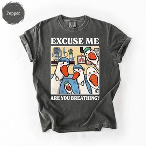 May include: Dark gray t-shirt with a humorous graphic featuring cartoon geese dressed as medical professionals. The text reads "EXCUSE ME ARE YOU BREATHING?" The geese are wearing stethoscopes and medical caps.
