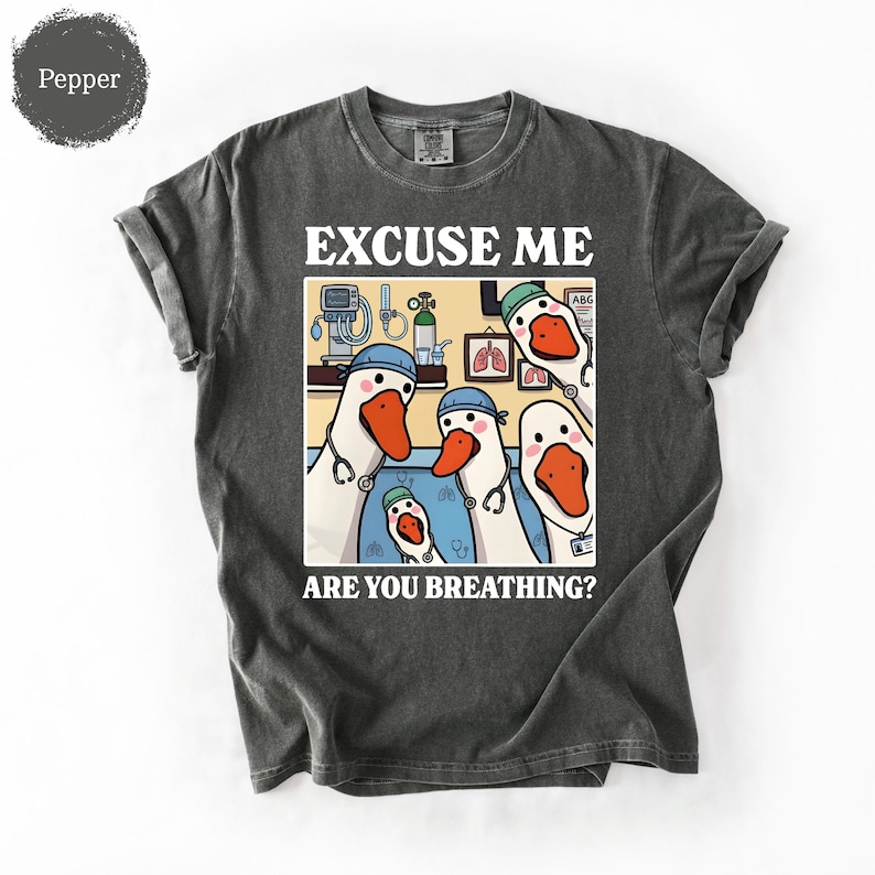 May include: Dark gray t-shirt with a humorous graphic featuring cartoon geese dressed as medical professionals. The text reads "EXCUSE ME ARE YOU BREATHING?" The geese are wearing stethoscopes and medical caps.