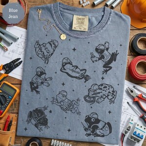 May include: A blue jean colored t-shirt featuring a repeating black graphic print of various frogs wearing construction hard hats and utility vests, some holding tools like pliers and screwdrivers, interspersed with small four-pointed star shapes.