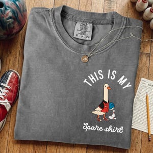 May include: Gray Comfort Colors t-shirt with a bowling goose graphic. The goose wears a red and black jacket and holds a bowling ball. Text reads "THIS IS MY Spare shirt". A bowling shoe and ball are visible.