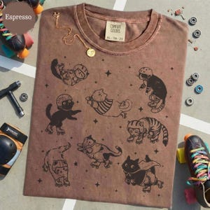 May include: Espresso-coloured t-shirt featuring a graphic of cats on roller skates. The shirt has a Comfort Colors tag, XL size. A gold necklace with a pendant is draped on the shirt. Roller skates and skate parts are in the background.