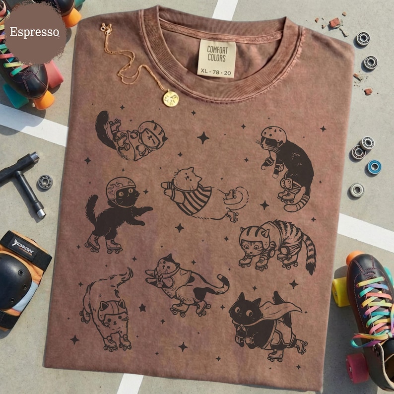 May include: Espresso-coloured t-shirt featuring a graphic of cats on roller skates. The shirt has a Comfort Colors tag, XL size. A gold necklace with a pendant is draped on the shirt. Roller skates and skate parts are in the background.