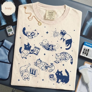 May include: Ivory-colored t-shirt featuring a navy blue illustration of cats in a radiology theme. The cats are depicted with x-rays, stethoscopes, and other medical equipment. The shirt has a crew neck and a Comfort Colors tag.