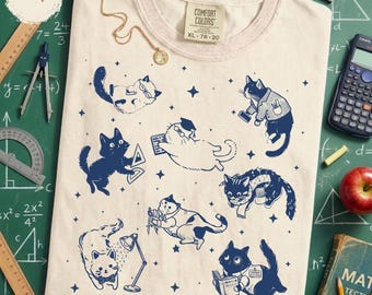 Könnte beinhalten: Elfenbeinfarbenes T-Shirt mit einem marineblauen Katzenmotiv. Die Katzen sind in verschiedenen Posen dargestellt, einige tragen Brillen oder halten Bücher und geometrische Werkzeuge. Das Shirt hat den Text "COMFORT COLORS XL 78-20".