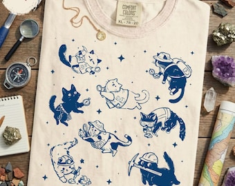 May include: An ivory t-shirt featuring a navy blue graphic print of various cats dressed as explorers and geologists with gear like hats, vests, and pickaxes. The shirt has a Comfort Colors label at the neck with the text XL 78-20.