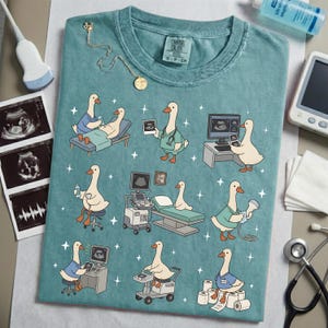 May include: Teal-colored t-shirt featuring cartoon geese in medical settings, including ultrasound scans and patient care. The design includes medical equipment and star accents. A gold necklace and ultrasound images are also visible.