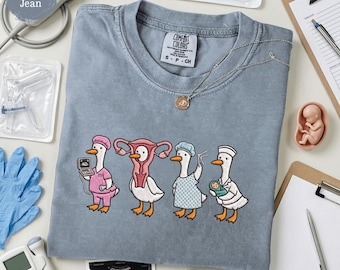May include: A blue jean colored t-shirt featuring a graphic of four white ducks dressed in medical attire. One duck wears a pink scrub, one has a uterus-shaped head, one wears a surgical cap and gown, and one holds a newborn baby.