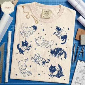 May include: Ivory-colored Comfort Colors t-shirt, size XL, featuring a navy blue graphic of cats in construction-themed poses. The cats are depicted with blueprints, tools, and hard hats. A gold necklace is draped on the shirt.
