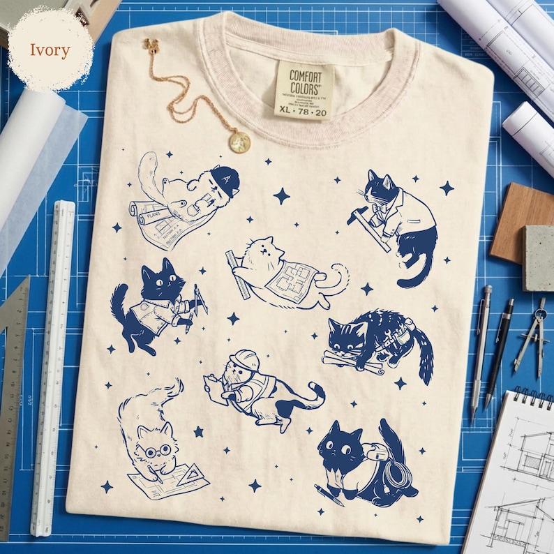 May include: Ivory-colored Comfort Colors t-shirt, size XL, featuring a navy blue graphic of cats in construction-themed poses. The cats are depicted with blueprints, tools, and hard hats. A gold necklace is draped on the shirt.