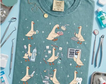 Könnte beinhalten: Ein meergrünes T-Shirt mit einem Grafikdruck von Zeichentrickgänsen, die verschiedene zahnärztliche Aufgaben ausführen, darunter das Halten von Zahnbürsten, Zahnpasta, Zahnarztinstrumenten und Röntgenbildern. Comfort Colors Etikett am Kragen.