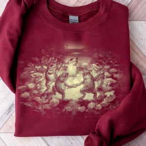 May include: Burgundy sweatshirt featuring a vintage-style illustration of dancing frogs in a pond setting. The frogs are depicted in a circle, holding hands, with a soft, sepia-toned color palette. The sweatshirt has a crew neck and long sleeves.
