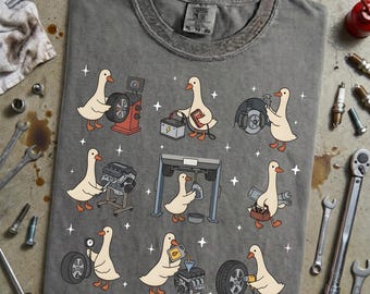 Könnte beinhalten: Graues T-Shirt mit einem Cartoon-Design einer Gans, die an Autoteilen arbeitet. Die Gans wird mit Werkzeugen und Autoteilen dargestellt, darunter Reifen, Motoren und eine Batterie. Das Shirt hat einen Rundhalsausschnitt und kurze Ärmel.