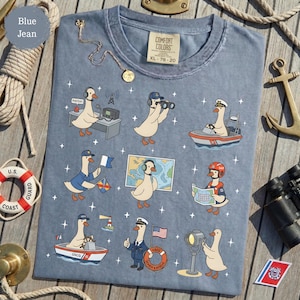 May include: Blue jean Comfort Colors t-shirt featuring cartoon geese in various Coast Guard roles. The geese are depicted operating a radio, using binoculars, navigating a boat, and more. The shirt has a tag that reads XL 78-20.
