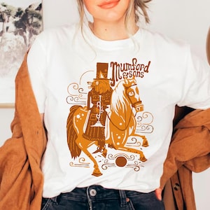 May include: White t-shirt featuring a whimsical illustration of a figure playing a trumpet while riding a horse. The design is in shades of brown and orange, with the text "Mumford & Sons" in a decorative font.