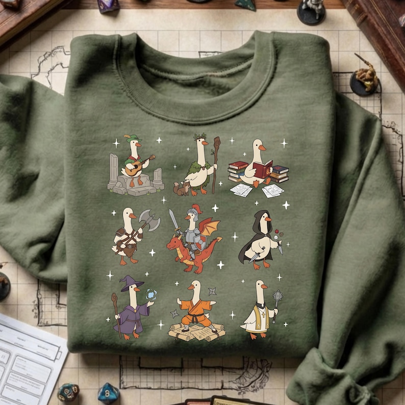 May include: Green sweatshirt featuring a whimsical design of geese in various fantasy roles. The geese are depicted as a bard, a cleric, a knight, and a wizard, among others. The design is detailed with stars and other decorative elements.