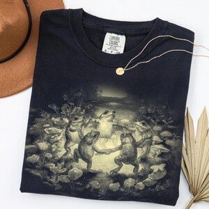 May include: Dark navy blue t-shirt featuring a whimsical illustration of dancing frogs in a forest setting. The graphic is in shades of gray and cream. The shirt has a crew neck and a tag that says "Comfort Colors". A gold necklace is draped across the shirt.