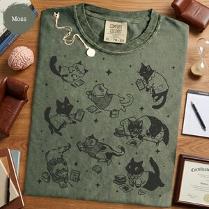 May include: A moss green t-shirt featuring a black graphic print of various cats in professional attire, including cats wearing glasses, sweaters, and lab coats, surrounded by small star icons and books, with a Comfort Colors label at the neck.