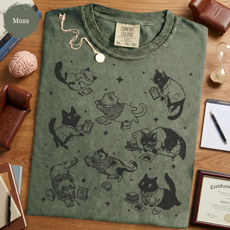 May include: A moss green t-shirt featuring a black graphic print of various cats in professional attire, including cats wearing glasses, sweaters, and lab coats, surrounded by small star icons and books, with a Comfort Colors label at the neck.