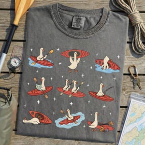 May include: Dark gray t-shirt featuring a whimsical design of cartoon geese kayaking in red kayaks. The geese are depicted in various kayaking scenarios, with paddles, maps, and fishing rods. The shirt has a crew neck and short sleeves.