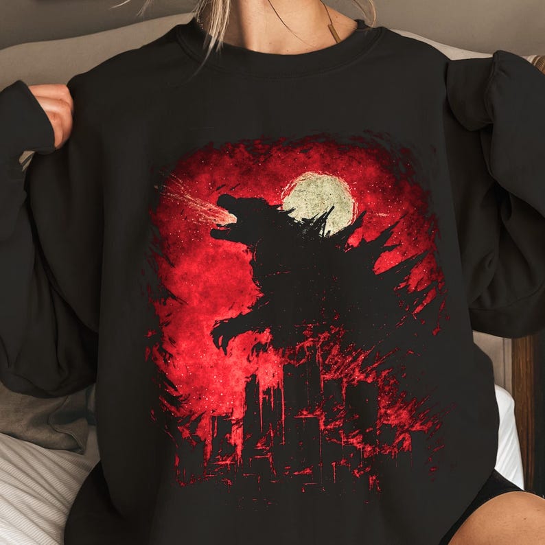 May include: Black sweatshirt featuring a Godzilla graphic. The design shows the silhouette of Godzilla against a red background with a full moon and city skyline. The monster is breathing fire, creating a dramatic scene.