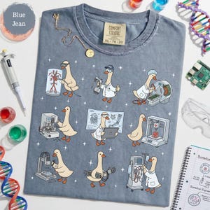 May include: Blue jean Comfort Colors t-shirt featuring a whimsical design of cartoon geese in lab coats, engaged in various scientific activities. The geese are depicted with medical equipment and technology. The shirt is size XL, with measurements 78-20.