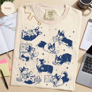 May include: An ivory t-shirt featuring a dark blue graphic print of multiple Corgi dogs in professional office attire. The dogs are depicted working with laptops, tablets, and project charts. The shirt has a Comfort Colors brand neck label.