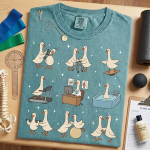 Pode incluir: Camiseta azul-petr&oacute;leo com gansos de desenho animado em v&aacute;rios cen&aacute;rios de fisioterapia. Os gansos s&atilde;o retratados usando uma esteira, prancha de equil&iacute;brio e outros equipamentos de reabilita&ccedil;&atilde;o. Uma pequena etiqueta na camisa diz "Comfort Colors". Um colar de ouro est&aacute; sobre a camisa.