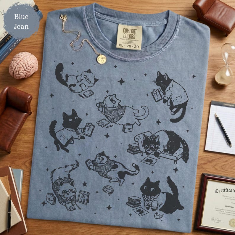 May include: A blue jean colored t-shirt featuring a black graphic print of various cats in professional attire, including cats with glasses, briefcases, and clipboards, scattered among small star shapes. The shirt has a Comfort Colors brand label.