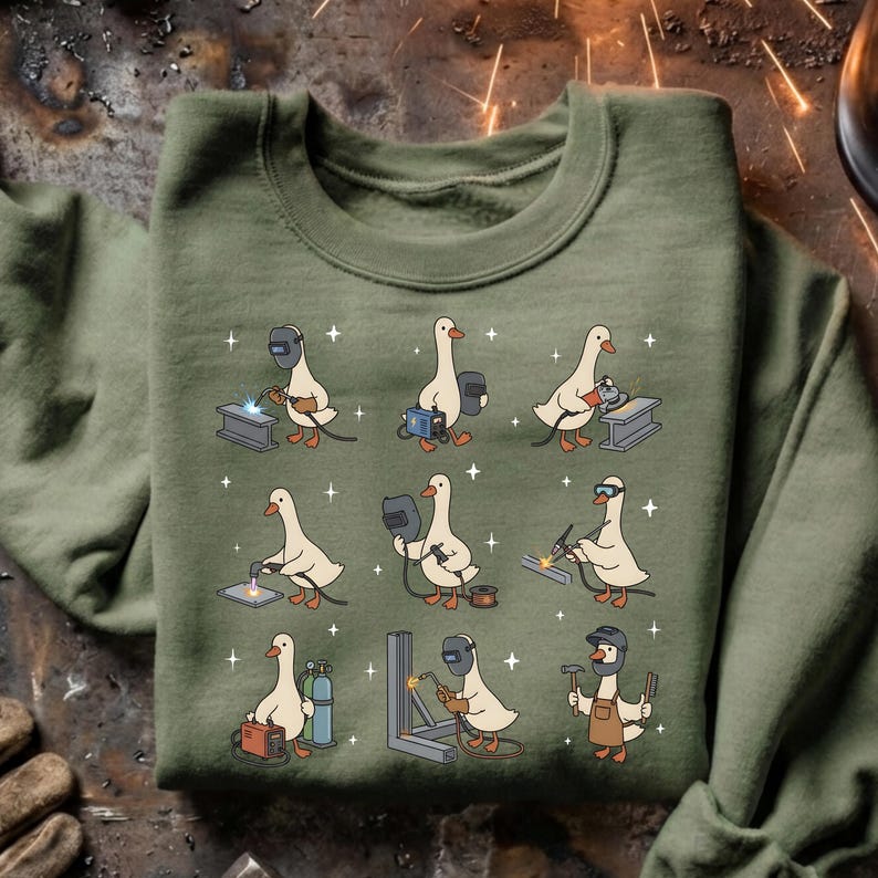 May include: Olive green sweatshirt featuring a repeating pattern of cartoon geese welding. Each goose is depicted with welding gear, working on various metal structures. The design includes sparks and a playful, whimsical style.