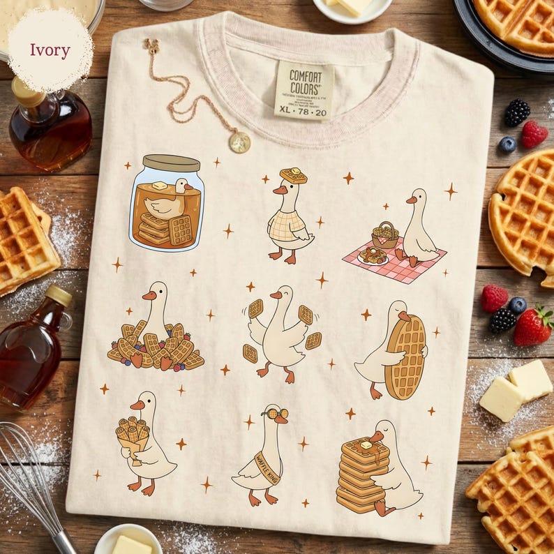 May include: Ivory-colored t-shirt with a Comfort Colors tag, featuring cartoon geese with waffles. The geese are depicted in various scenes, including holding waffles, in a jar, and on a picnic blanket. The shirt has a round neckline.