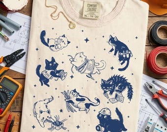 Puede incluir: Camiseta Comfort Colors color marfil con ilustraciones de gatos azul marino. Los gatos están representados con herramientas y equipos eléctricos. La camiseta tiene cuello redondo y una etiqueta que dice XL 78-20.