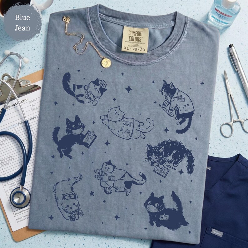 May include: Blue jean Comfort Colors t-shirt featuring a pattern of cartoon cats in medical roles. The cats are depicted as doctors, nurses, and wearing face masks. A gold necklace and medical equipment are also visible.