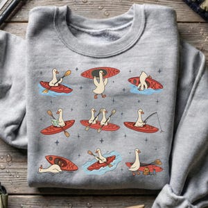 May include: Gray sweatshirt featuring a whimsical design of cartoon geese kayaking, fishing, and carrying kayaks. The geese are depicted in red kayaks with paddles, maps, and fishing rods. The design is repeated across the sweatshirt.