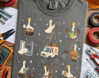May include: Grey t-shirt with a cartoon design of geese in yellow hard hats working as electricians. The geese are shown working on electrical panels, wiring, and transformers. The shirt has a crew neck and short sleeves.