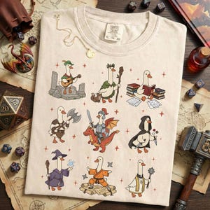 May include: Cream-colored t-shirt featuring cartoon geese in various fantasy roles, such as a bard, a wizard, and a knight riding a dragon. The shirt has the text "Comfort Colors XL 70-20" printed on the upper right side. The design includes red star accents.